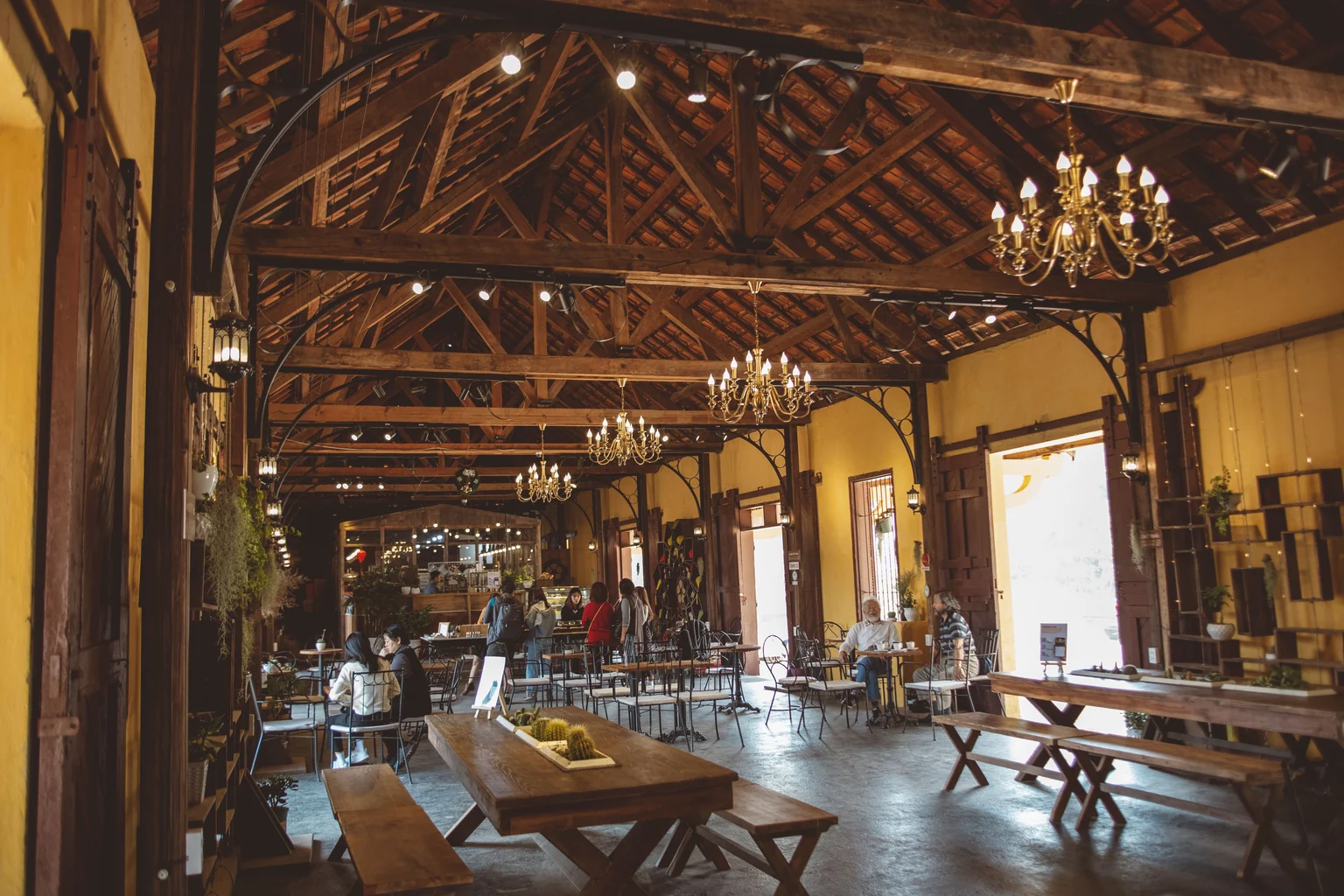 Interior de café com vigas de madeira, mesas, lustres e pessoas ao fundo