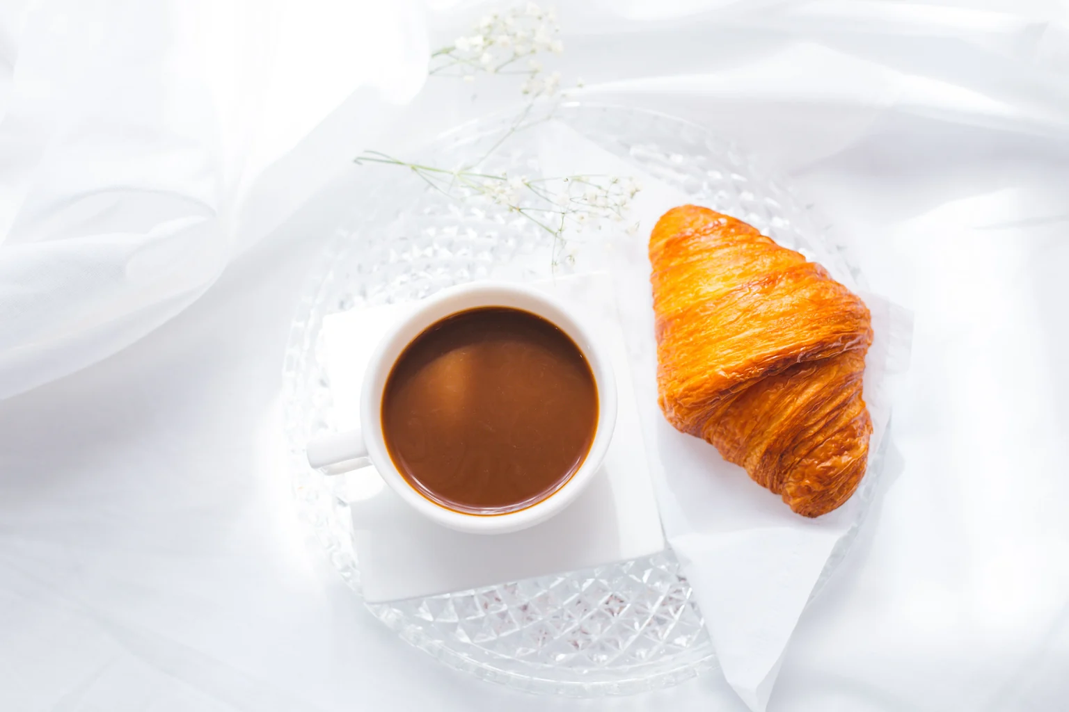 Xícara de café e croissant sobre um prato de vidro, em uma mesa com toalha clara